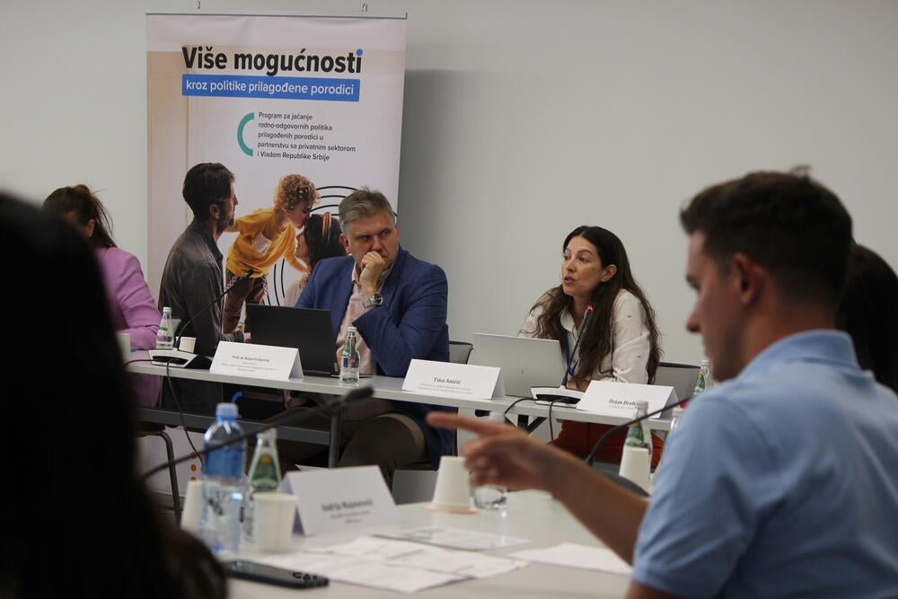 Žene govore na konferenciji, a muškarac pokazuje na nešto na ekranu. Ljudi u konferencijskoj sali slušaju govornika.