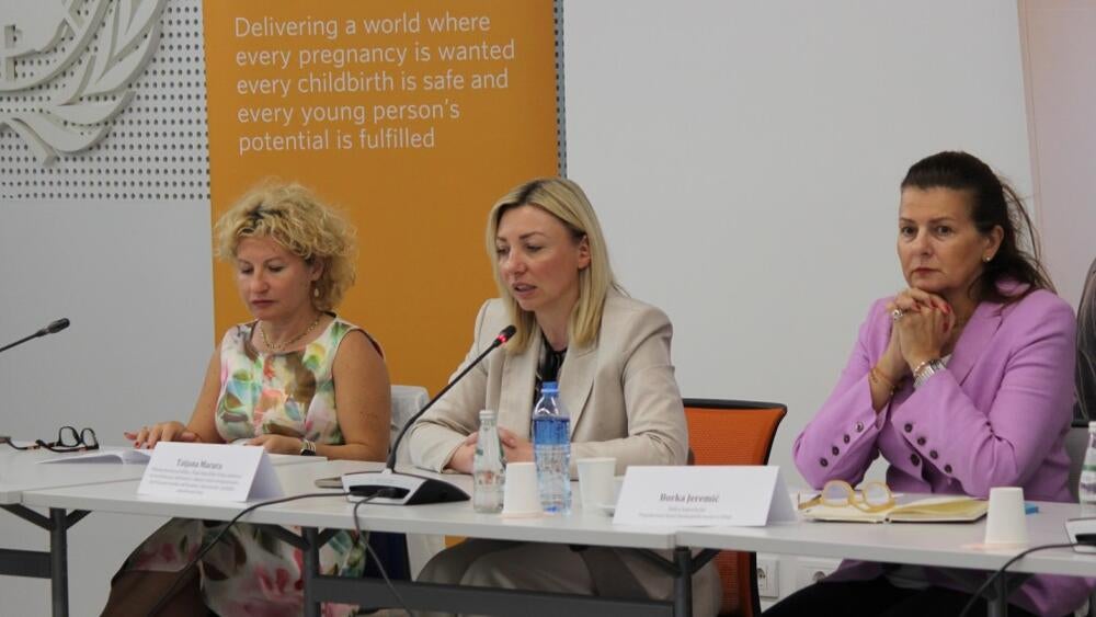 Tri žene sede za konferencijskim stolom i slušaju govornika.
