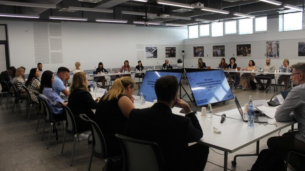 Vidljiva je konferencijska sala, ljudi sede i slušaju govornika. U centru konferencijske sale nalaze se četiri TV ekrana na kojima se prikazuje prezentacija.